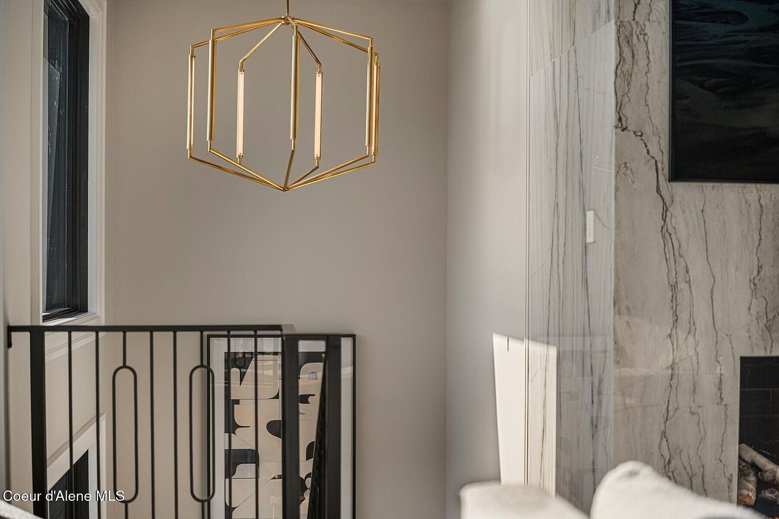 This interior shot showcases a modern hallway or staircase landing featuring a striking gold geometric chandelier. The space is decorated with a black metal railing, artwork, and a marble-clad wall with a fireplace, creating a sophisticated and stylish transition area within the home. Natural light streams in from a nearby window, enhancing the neutral color palette.