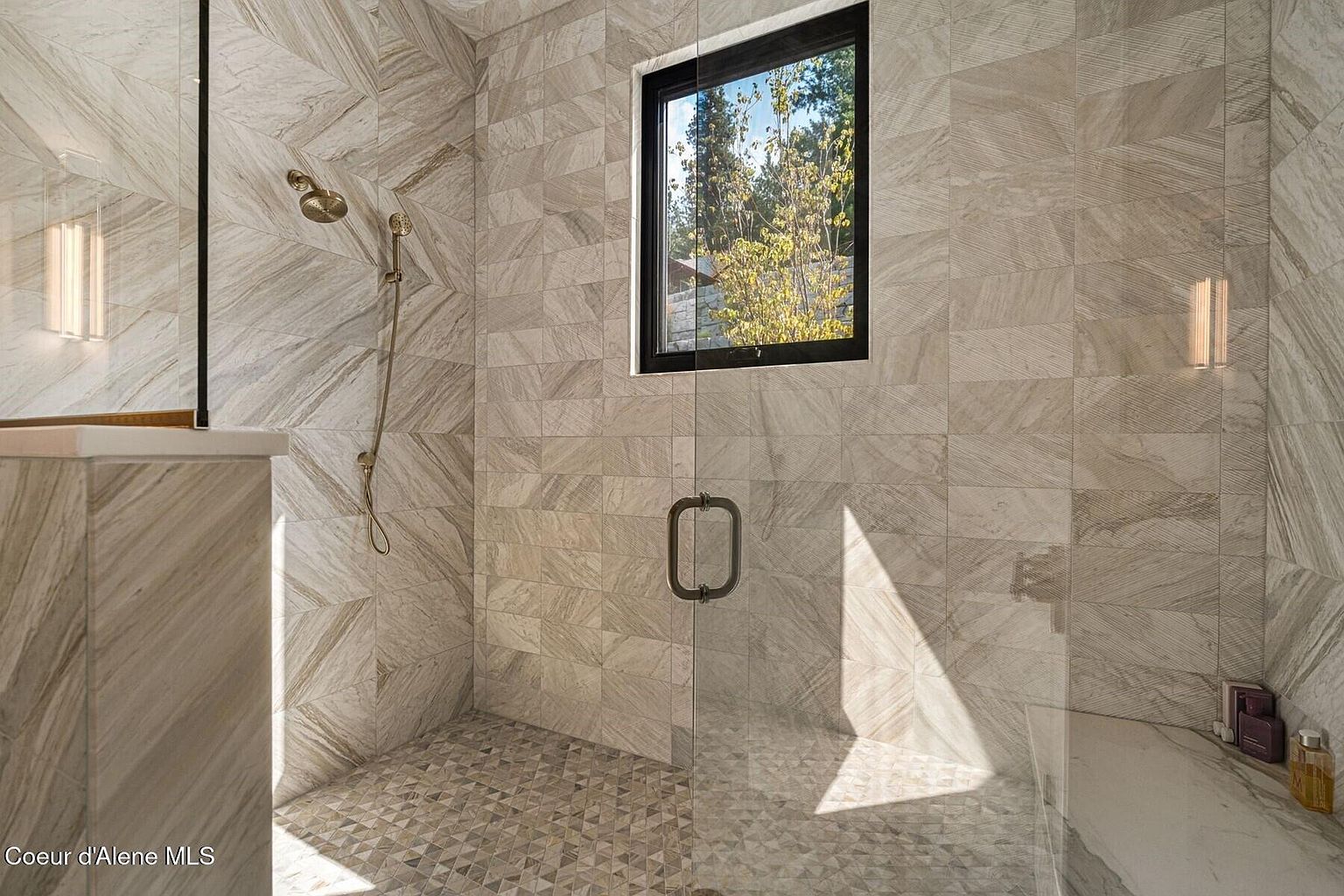 This is a luxurious primary bathroom featuring a spacious walk-in shower with glass doors and walls. The shower is tiled with a light-colored stone in a linear pattern, complemented by a mosaic tile floor. A window provides natural light, and the overall design exudes a modern and spa-like ambiance.