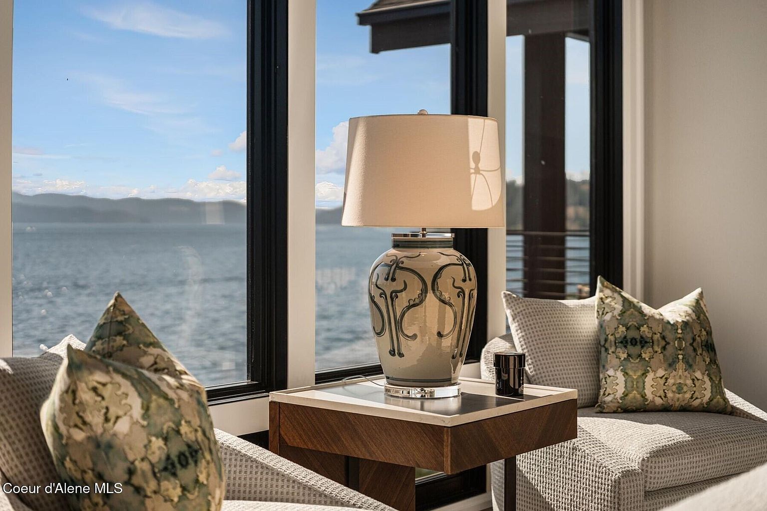 This interior shot showcases a cozy living space with a focus on natural light and serene views. A stylish lamp sits on a side table next to a comfortable seating area with decorative pillows. The large window frames a picturesque lake and mountain landscape, creating a tranquil and inviting atmosphere.