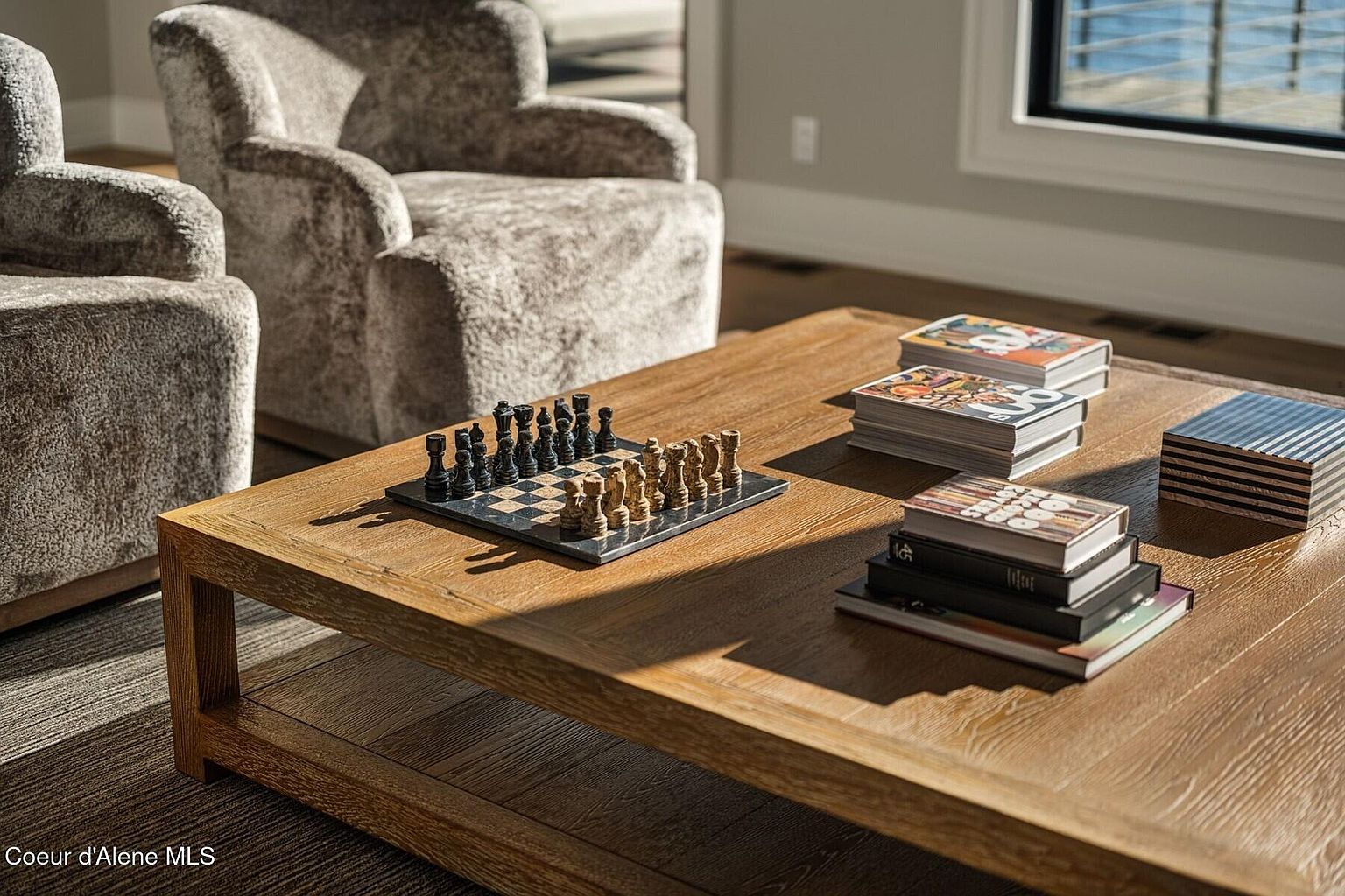 This image showcases a cozy living room interior, focusing on a wooden coffee table adorned with a chess set, books, and decorative objects. Two plush, textured armchairs are partially visible in the background, contributing to the room's inviting atmosphere. The natural light streaming in from a nearby window enhances the warm tones of the wood and the soft textures of the furniture, creating a comfortable and stylish living space.