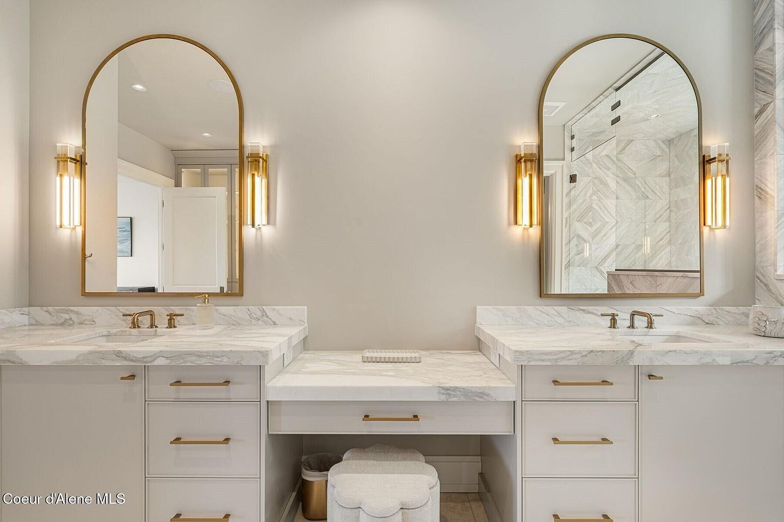 This primary bathroom features dual vanities with white cabinetry, marble countertops, and gold hardware. Arched mirrors are flanked by sconce lighting, creating a luxurious and well-lit space. A vanity area with a stool is situated between the sinks, adding to the room's functionality and elegance.