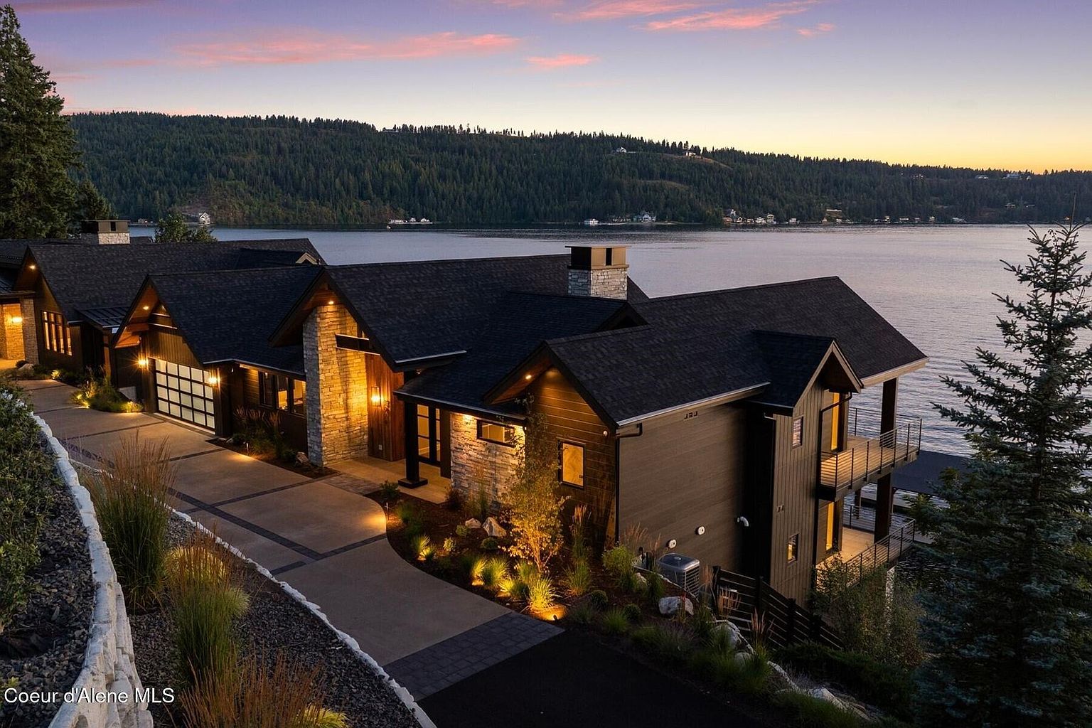 This is a stunning front view of a modern luxury home situated near a lake. The house features a combination of dark wood siding and stone accents, with a dark roof and well-lit landscaping. A paved driveway leads to a garage, and the overall impression is one of high-end elegance and serene lakeside living.