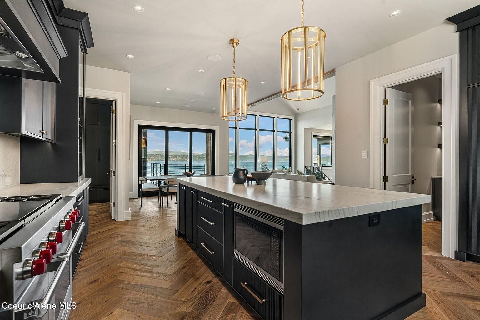 This is a view of a modern kitchen featuring a large island with a marble countertop and dark cabinetry. The kitchen is open to a living area with large windows offering a scenic view of the water. The flooring is a herringbone pattern hardwood, and the lighting includes pendant fixtures above the island.