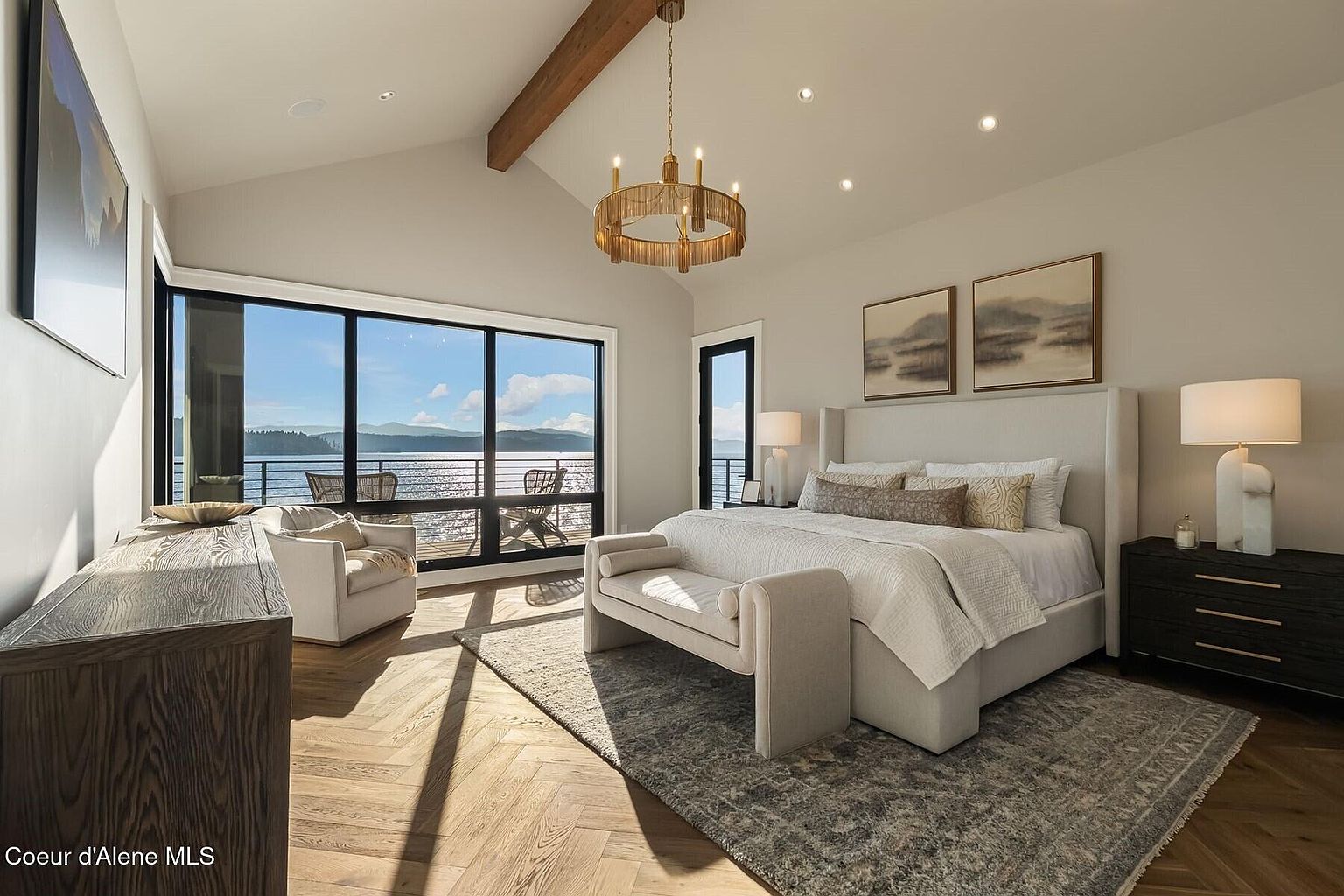 This is a luxurious primary bedroom featuring a large bed with a padded headboard, a bench at the foot of the bed, and a dark wood nightstand with a lamp. The room has large windows offering a stunning view of the water and mountains, and a chandelier hangs from the vaulted ceiling. The overall style is elegant and serene, creating a relaxing atmosphere.