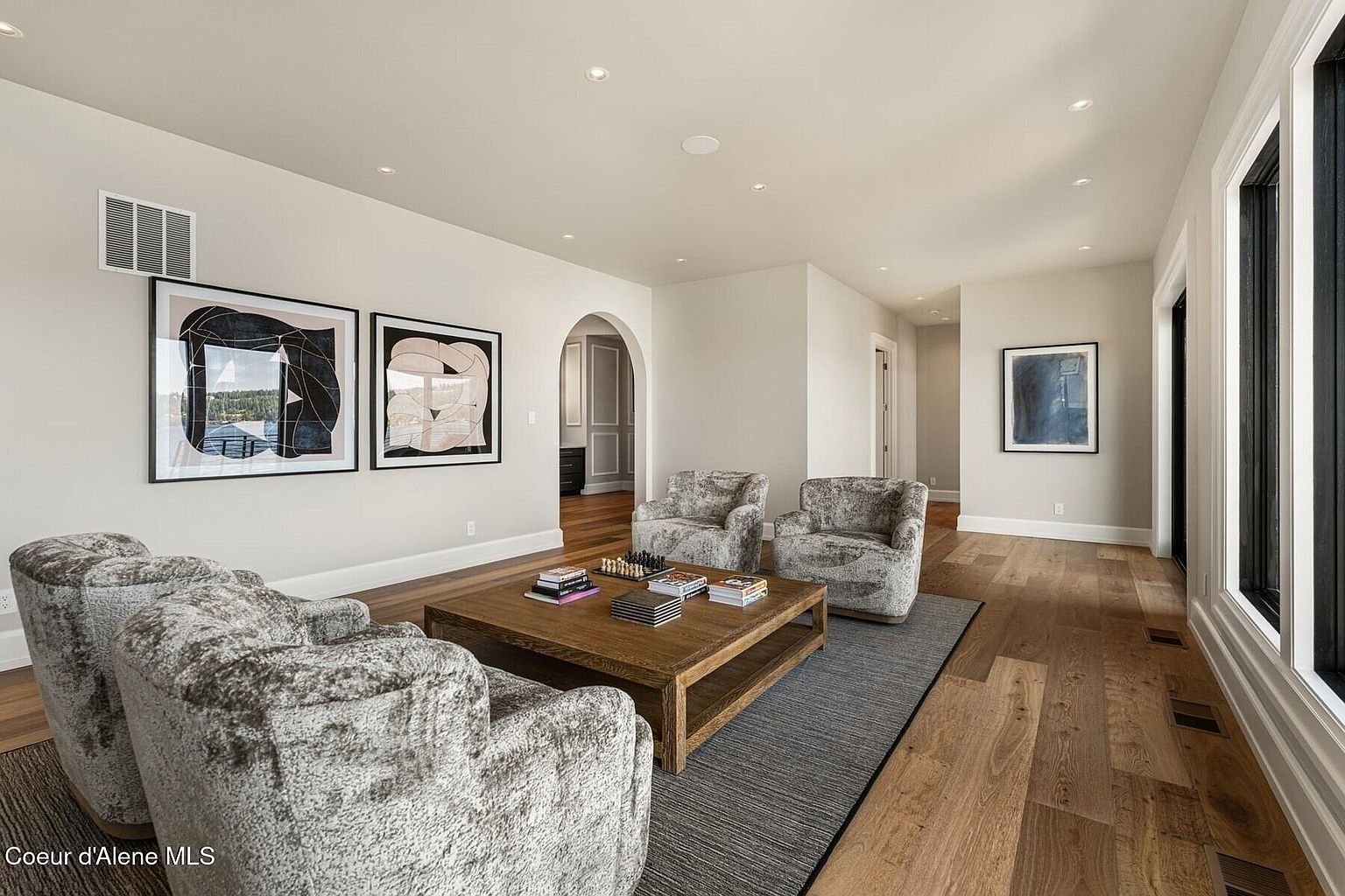 This living room features a neutral color palette with hardwood floors and a gray area rug. The room is furnished with plush, gray armchairs and a wooden coffee table adorned with books and a chess set. Artwork hangs on the walls, and natural light streams in through the windows, creating a warm and inviting atmosphere.