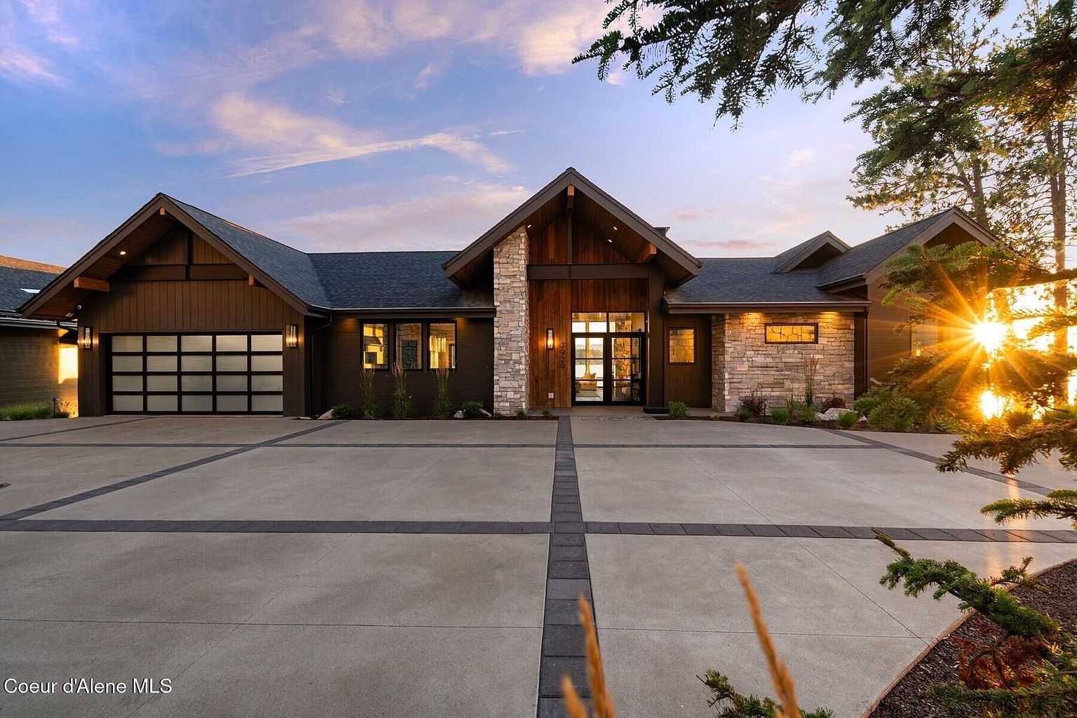 This is a front exterior view of a modern luxury home. The house features a combination of wood siding and stone accents, with a prominent gabled entrance. A large concrete driveway with decorative inlays leads to a multi-car garage, and the landscaping is well-maintained, creating a welcoming and upscale impression.