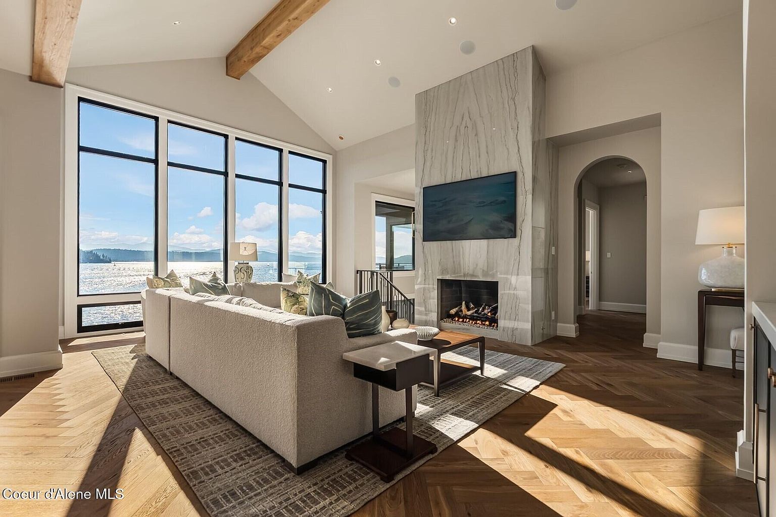 This is a bright and airy living room featuring a large window with a scenic lake view. The room is furnished with a comfortable sofa, a modern fireplace with a marble surround, and stylish decor. The hardwood floors and neutral color palette create a sophisticated and inviting atmosphere.