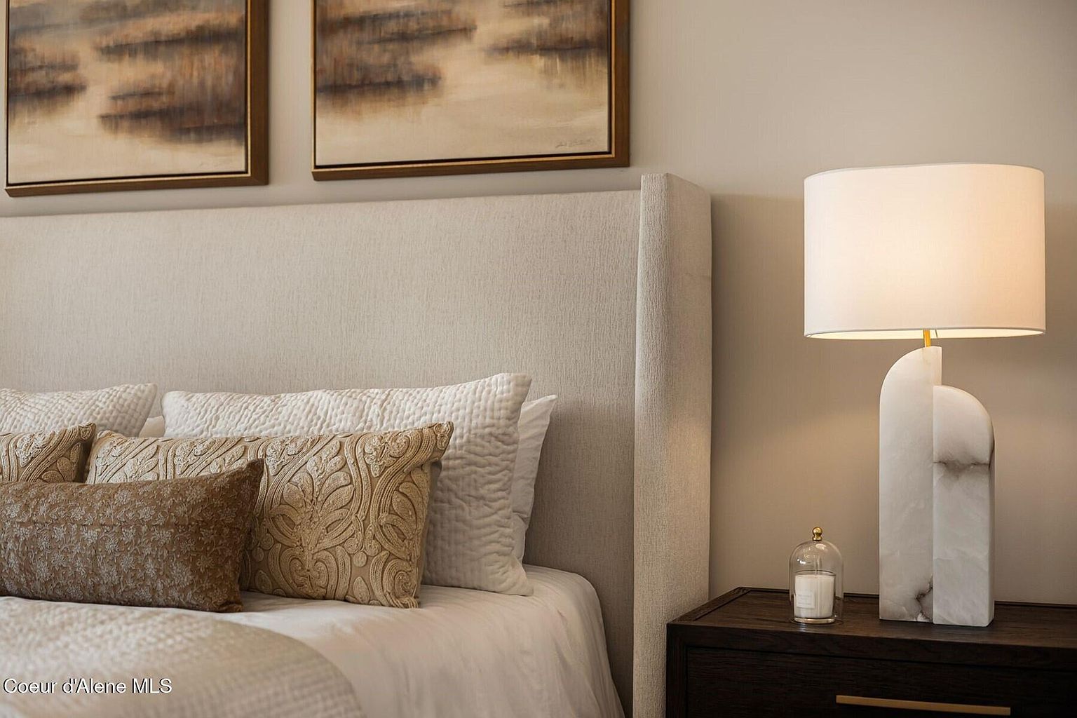 This is a well-staged primary bedroom featuring a large upholstered headboard, decorative pillows, and a bedside table with a modern lamp. The neutral color palette creates a serene and inviting atmosphere. The room is designed to showcase comfort and style, appealing to potential buyers looking for a relaxing retreat.