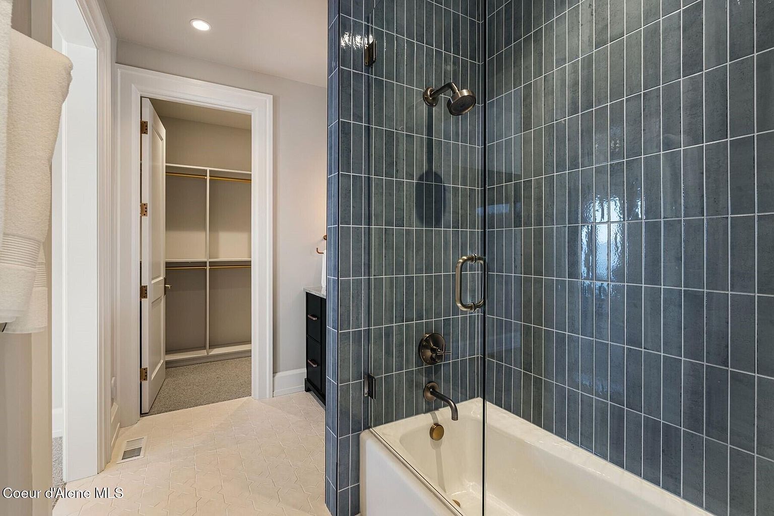 This bathroom features a modern shower-tub combination with blue vertical tiles and glass enclosure. The floor is tiled with a patterned design, and a walk-in closet is visible through an open doorway. The overall impression is clean and stylish, with a focus on contemporary design elements.