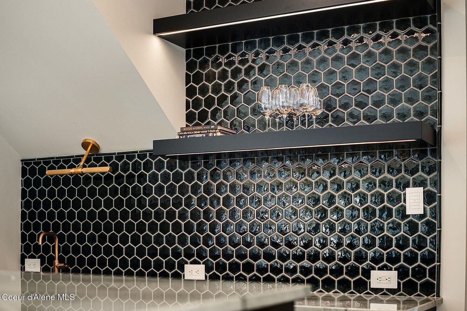 This image showcases a stylish kitchen backsplash featuring dark, hexagonal tiles with light grout, creating a striking geometric pattern. Two sleek, black shelves with integrated lighting add a modern touch, while a gold-toned faucet and electrical outlets provide functional accents. The overall impression is sophisticated and contemporary, highlighting the kitchen's attention to detail.