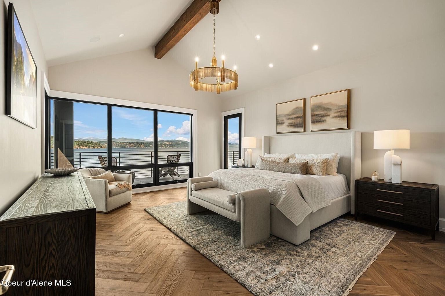 This is a luxurious primary bedroom featuring a king-size bed with a padded headboard, a bench at the foot of the bed, and a large window with a view of the water. The room is decorated in neutral tones with wood floors and a statement chandelier, creating a serene and upscale atmosphere. The room also features a dark wood dresser and two pieces of artwork above the bed.