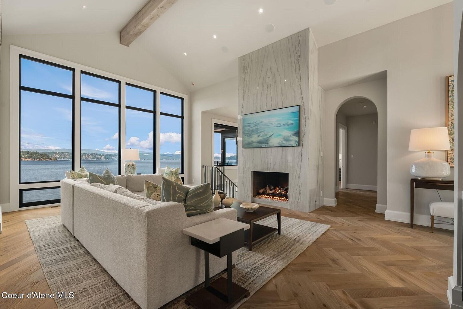 This is an interior shot of a luxurious living room featuring a large sectional sofa facing a modern fireplace with a mounted television. The room has expansive windows offering a scenic view of the water and sky. The flooring is hardwood laid in a herringbone pattern, and the overall aesthetic is bright, airy, and upscale.