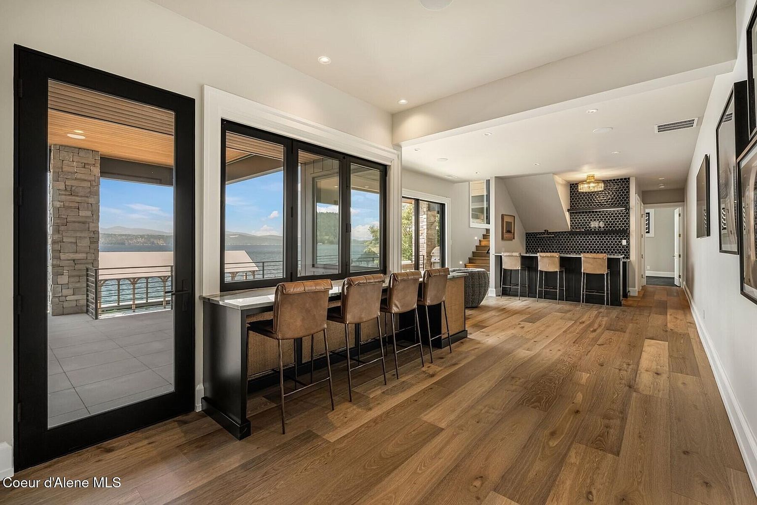 This interior shot showcases a modern living space with hardwood floors and a bar area featuring leather bar stools. Large windows offer a view of the water, and a doorway leads to an outdoor patio. The room also includes a staircase leading to a lower level with a bar and additional seating.