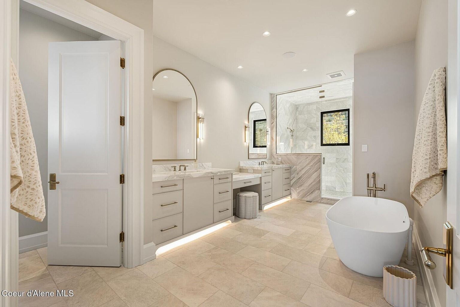 This is a luxurious primary bathroom featuring dual vanities with arched mirrors and elegant lighting fixtures. A freestanding bathtub sits near a glass-enclosed shower, and the neutral color palette creates a serene and spa-like atmosphere. The tile flooring and under-cabinet lighting add to the sophisticated design.