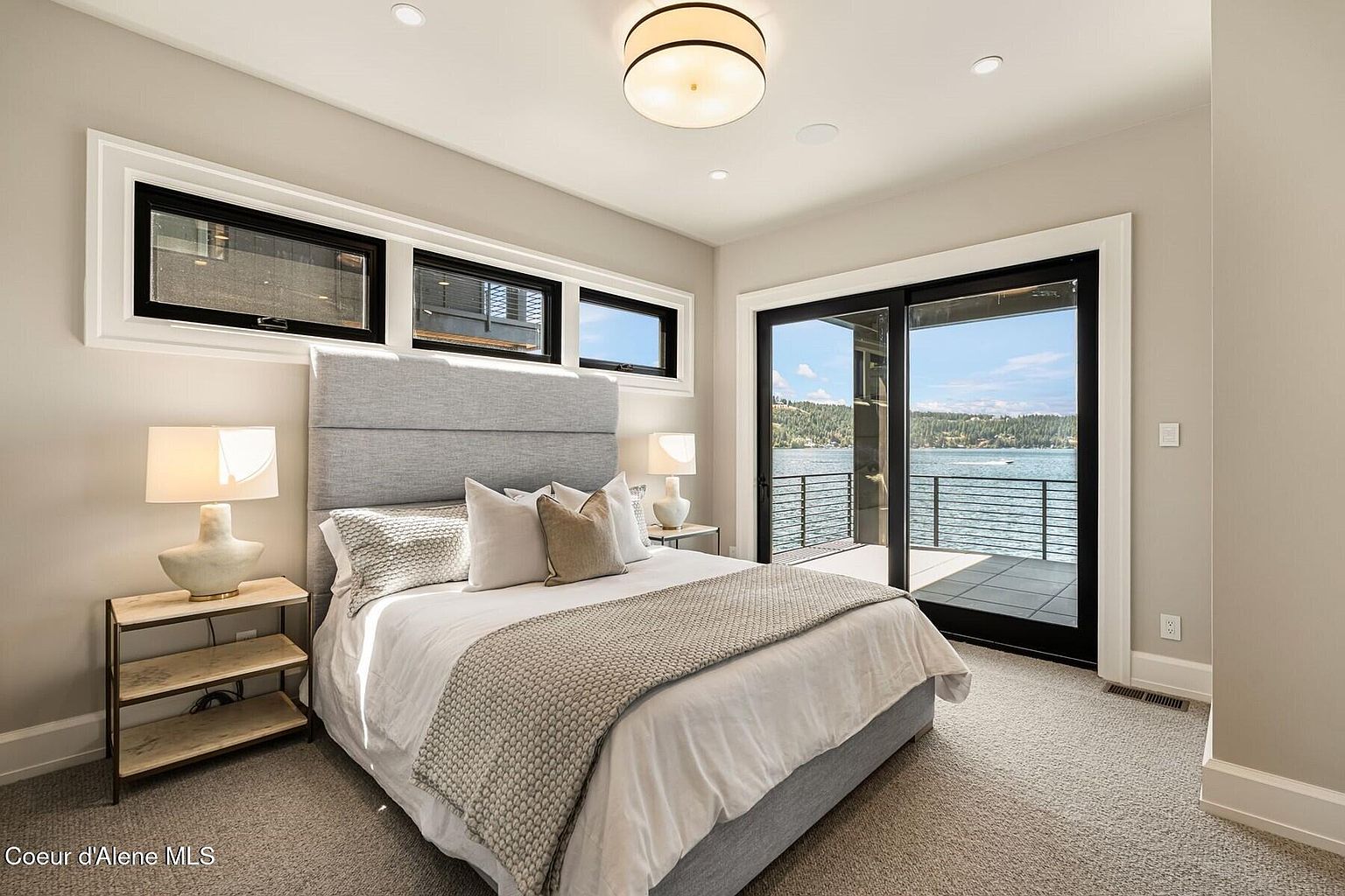 This is a bright and airy primary bedroom with a modern aesthetic. The room features a large bed with a gray upholstered headboard, neutral bedding, and a textured throw. Sliding glass doors lead to a balcony with a water view, and there are three horizontal windows above the bed, providing natural light.