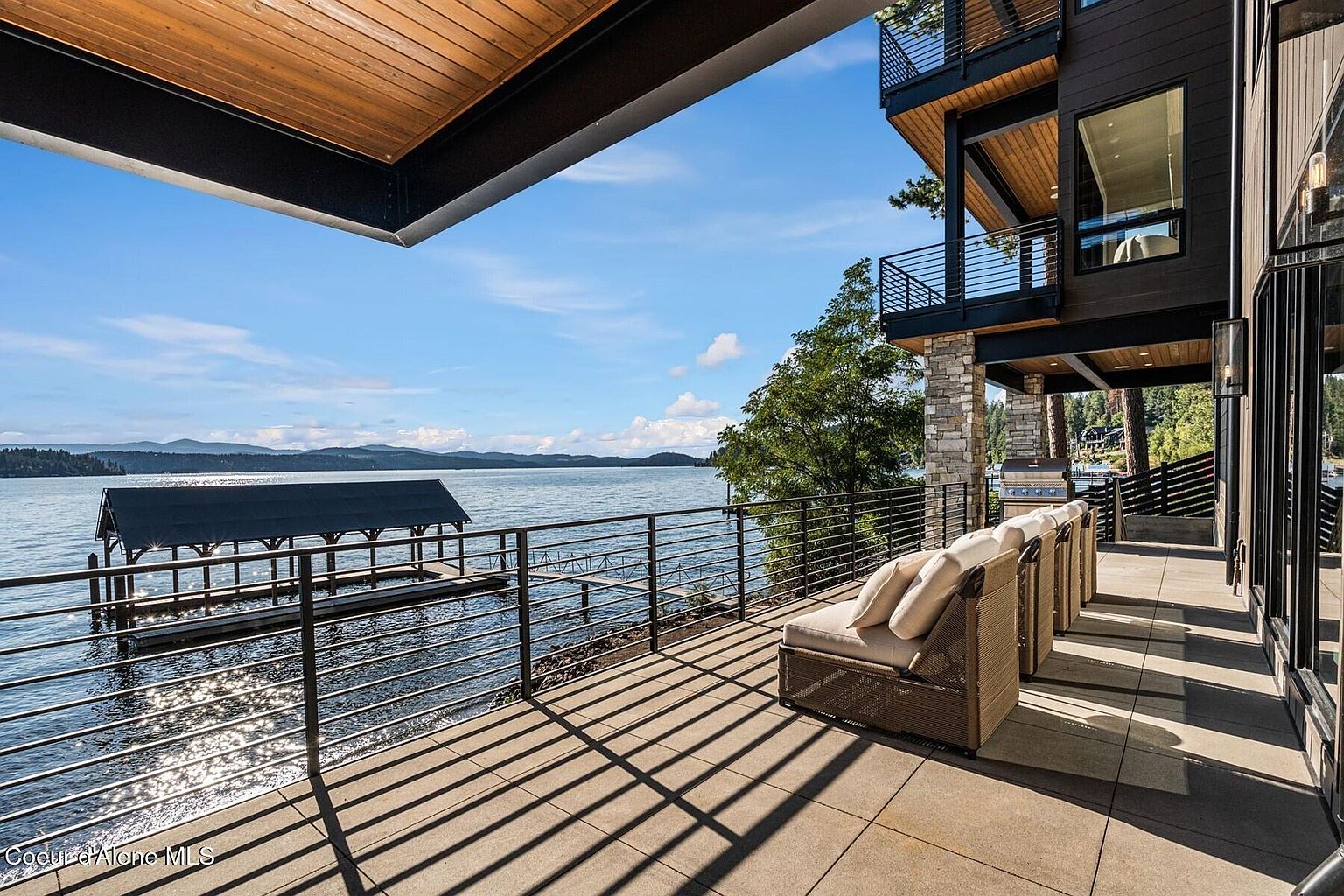 This image showcases a luxurious outdoor patio or deck area with stunning waterfront views. The space features comfortable lounge seating, a modern railing system, and a sleek, contemporary design that complements the adjacent building's architecture. The overall impression is one of high-end relaxation and sophisticated outdoor living.