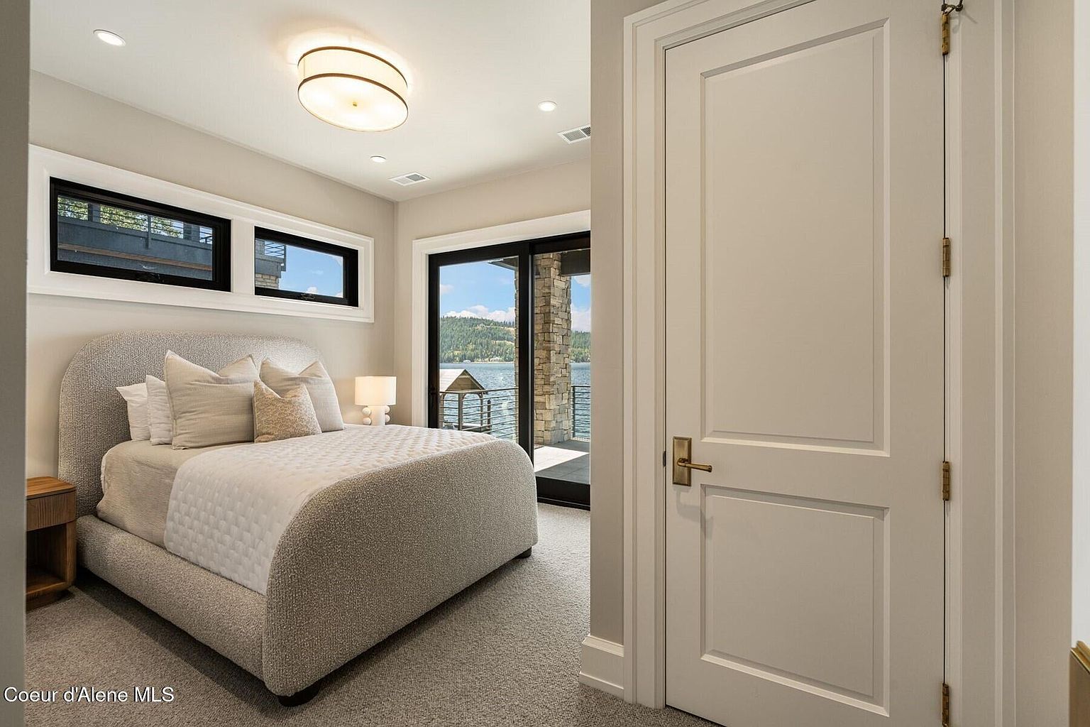 This is a primary bedroom featuring a large bed with a textured headboard and neutral-toned bedding. The room has two windows and a sliding glass door that leads to a balcony with a lake view. A white door with brass hardware is visible on the right, and the room is carpeted in a neutral color.