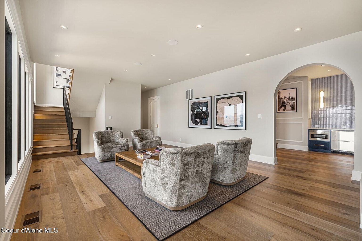 This is an interior shot of a living room featuring hardwood floors, a gray area rug, and four plush armchairs arranged around a wooden coffee table. Two framed abstract art pieces hang on the wall, and an arched doorway leads to another room with a small kitchen area. A staircase is visible on the left side of the image, adding architectural interest.
