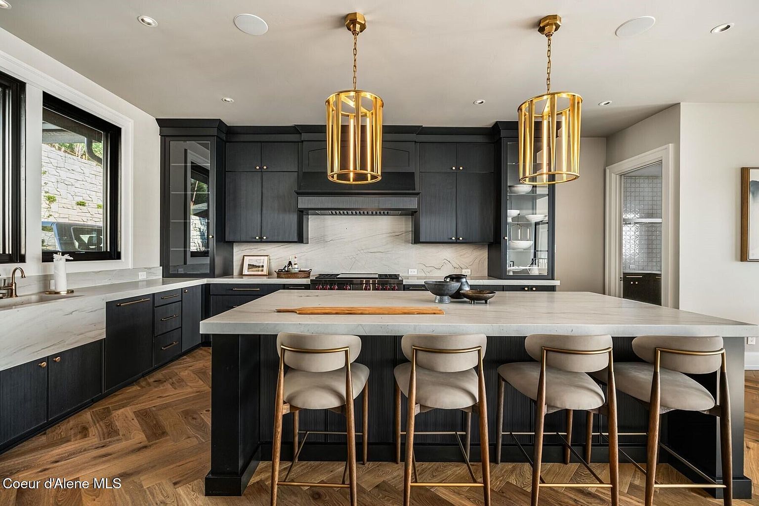 This is a well-lit kitchen featuring dark cabinetry, a marble backsplash, and a large island with seating. Two gold pendant lights hang above the island, adding a touch of elegance. The herringbone wood flooring complements the modern design, creating a sophisticated and inviting space.