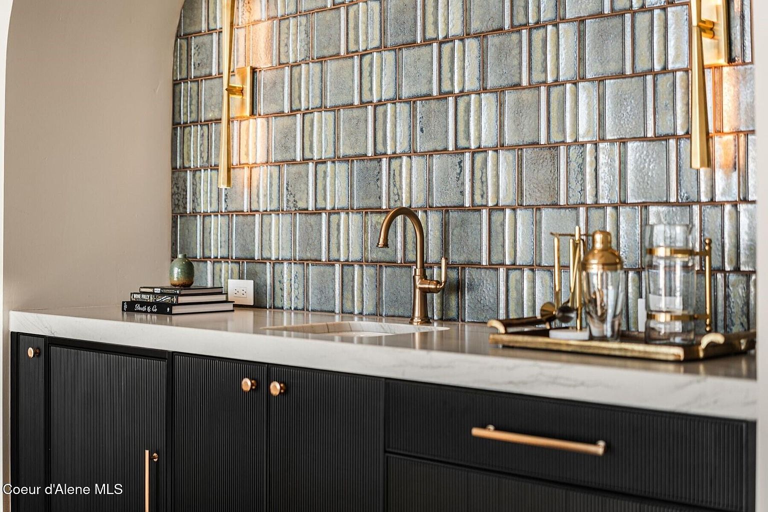 This image showcases a stylish bar area within a kitchen, featuring a textured tile backsplash in muted tones and elegant gold fixtures. The dark cabinetry with linear detailing and gold hardware adds a touch of sophistication, complemented by a light countertop. The scene is well-lit, highlighting the textures and finishes, creating an inviting and luxurious atmosphere.