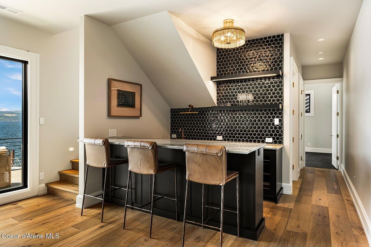This interior shot showcases a stylish wine cellar or bar area, featuring a black bar with a white countertop and three leather bar stools. The backdrop is adorned with black honeycomb tiles, complemented by floating shelves and a modern chandelier. The space is well-lit and exudes a sophisticated, contemporary vibe, perfect for entertaining.
