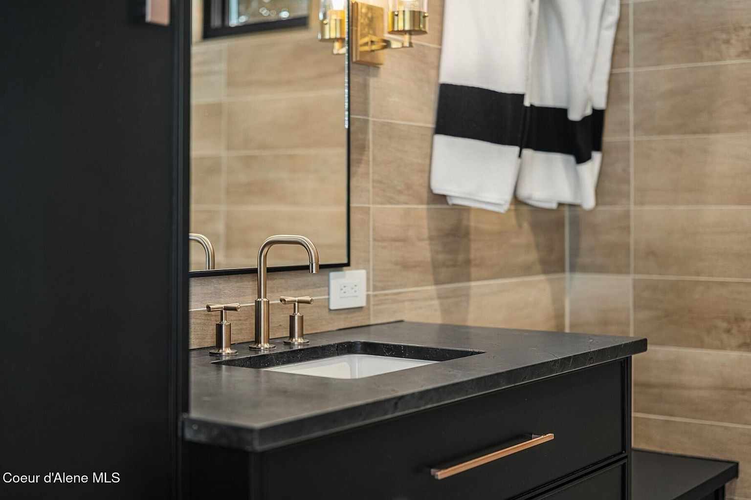 This is a well-appointed bathroom featuring a dark vanity with a stone countertop and a modern gold faucet. The walls are tiled in a neutral tone, and a black-framed mirror hangs above the sink. White towels with black stripes add a touch of elegance to the space.