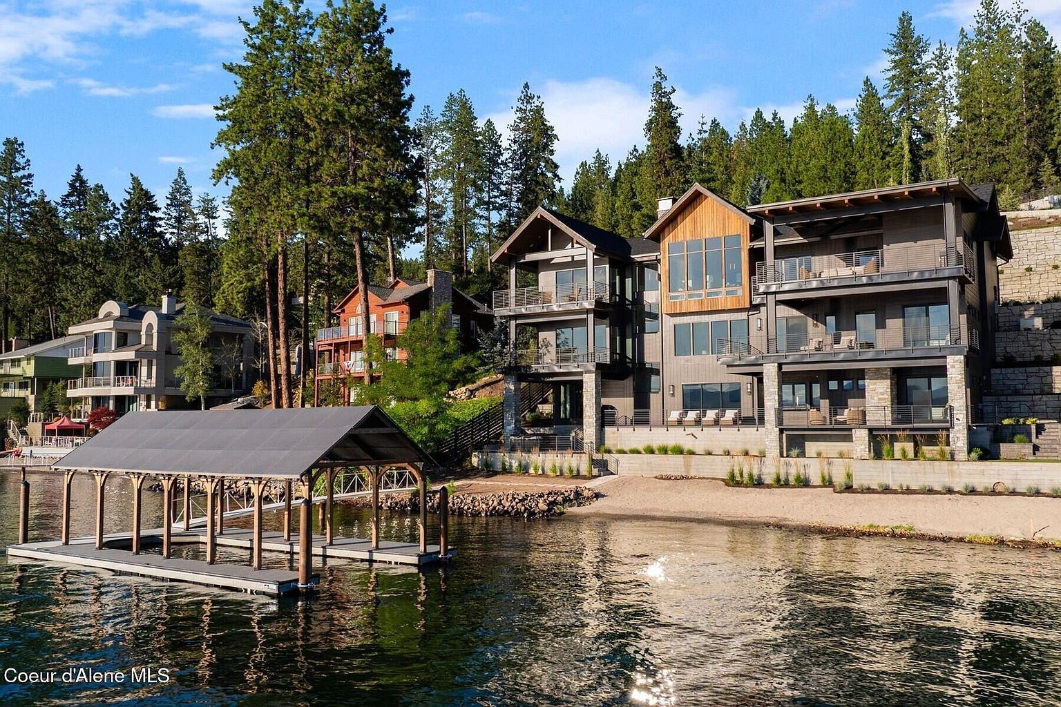 This image showcases the rear exterior of a luxurious lakefront home, featuring multiple levels with balconies and large windows that offer stunning views. The property includes a private beach area and a boat dock, blending seamlessly with the natural landscape of trees and water. The architectural style is modern and sophisticated, emphasizing outdoor living and waterfront access.