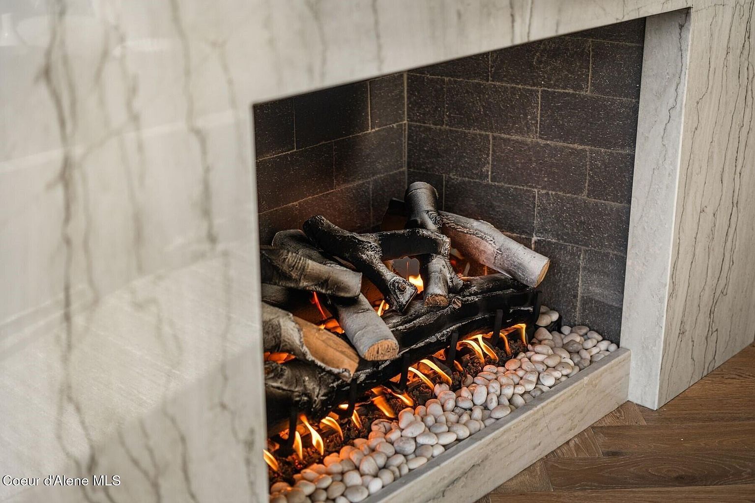 This image showcases a modern fireplace with a marble surround and a dark brick interior. The fireplace features realistic-looking logs and a bed of white pebbles, creating a cozy and inviting atmosphere. The design is sleek and contemporary, adding a touch of elegance to the space.