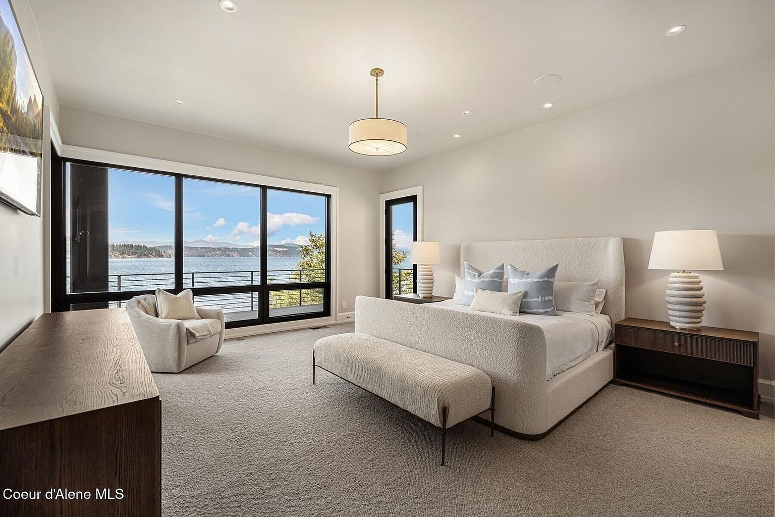 This is a bright and spacious primary bedroom featuring a large window with a scenic water view. The room is furnished with a plush bed, a bench, and a nightstand with a lamp. The neutral color palette and carpet create a serene and inviting atmosphere, perfect for relaxation.