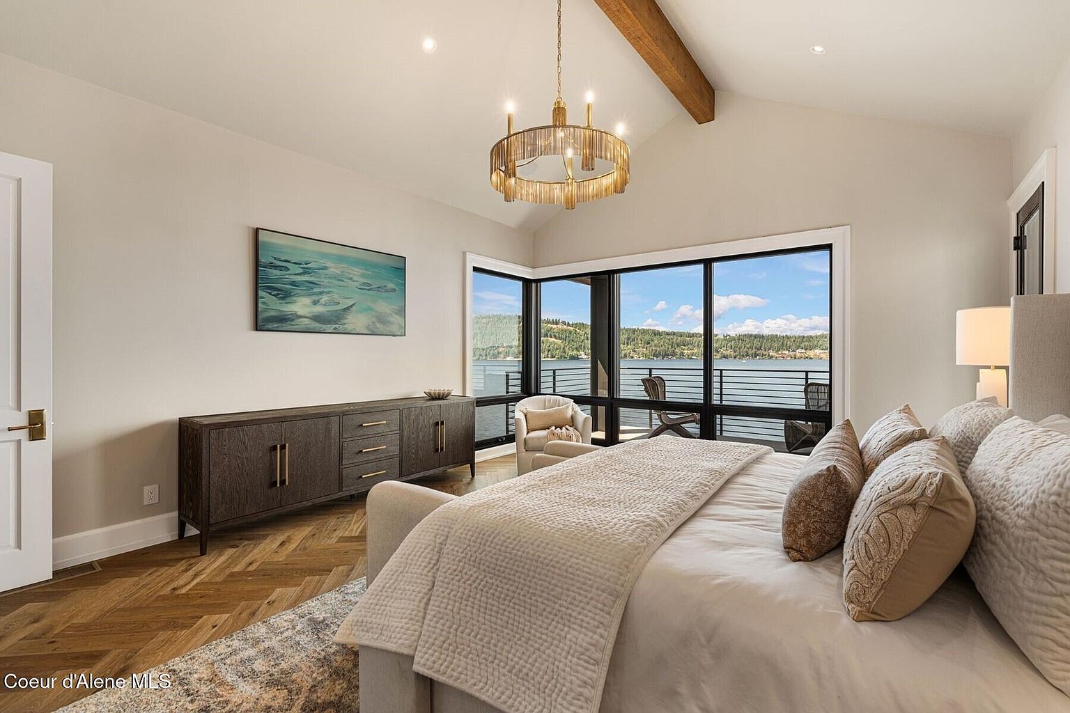 This is a luxurious primary bedroom featuring a king-size bed with plush pillows and a cozy throw blanket. The room boasts a stunning view of the lake through large windows, complemented by a stylish chandelier and a wooden beam on the ceiling. A dark wood dresser adds a touch of elegance, while the overall design creates a serene and inviting atmosphere.