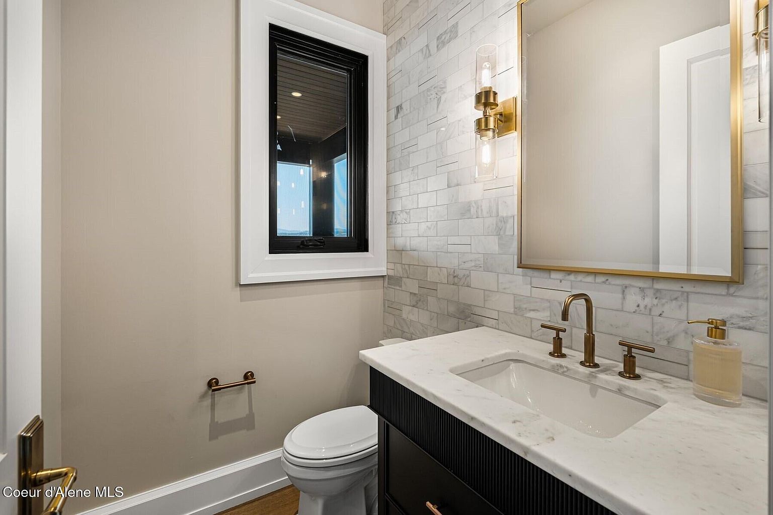This is a well-lit guest bathroom featuring a modern design. The vanity has a white marble countertop and a dark cabinet with vertical detailing. A rectangular mirror with a gold frame hangs above the sink, complemented by stylish wall-mounted light fixtures. The walls are partially covered with marble tiles, adding a touch of luxury.