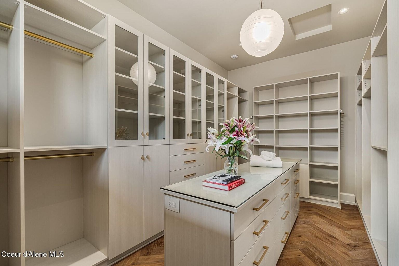 This is a well-organized walk-in closet featuring custom cabinetry with glass-fronted doors, drawers with gold hardware, and open shelving. A central island with a light countertop provides additional storage and display space, topped with flowers and books. The herringbone wood flooring adds a touch of elegance to the space.