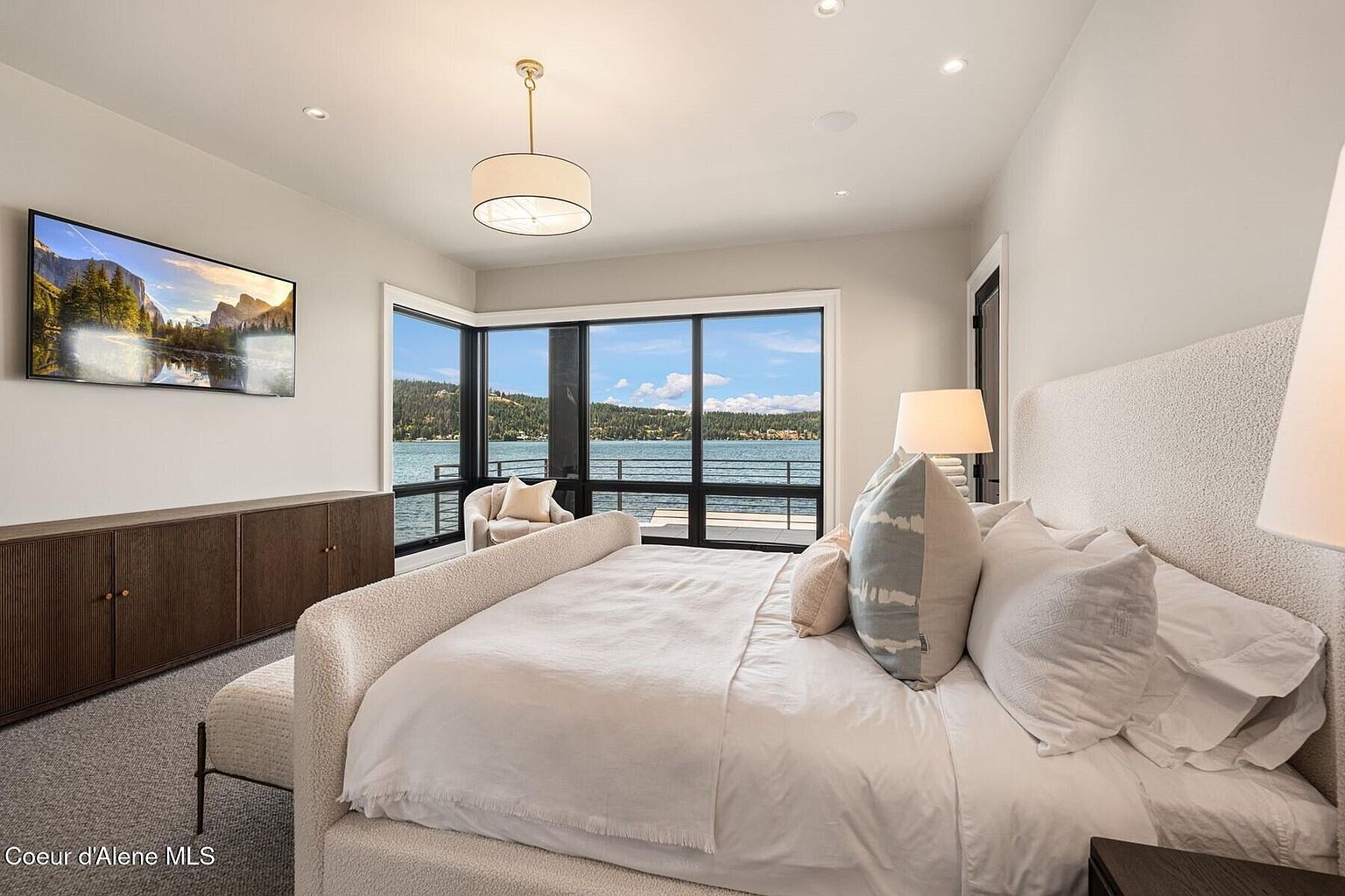 This is a bright and airy primary bedroom featuring a large bed with white linens and decorative pillows, a modern light fixture, and a wall-mounted television. Large windows offer a stunning view of the lake, and a built-in cabinet provides ample storage. The room exudes a sense of luxury and tranquility.