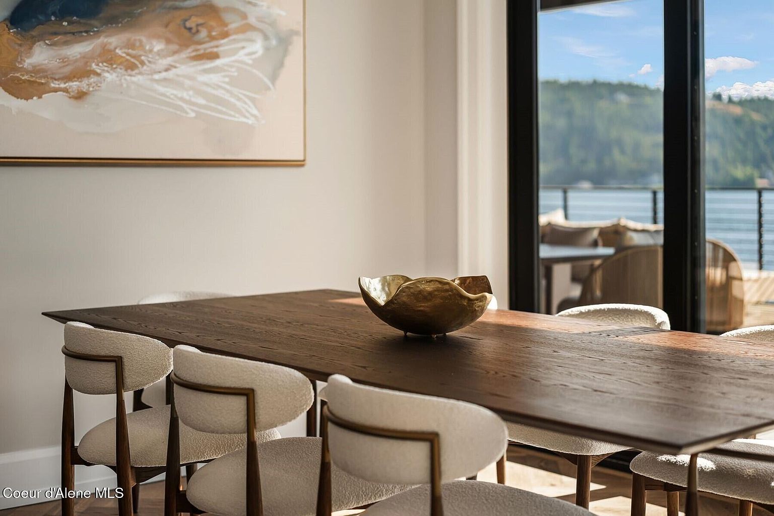 This image showcases a dining room with a long, dark wood table surrounded by modern chairs with white, textured upholstery and light wood frames. A decorative gold bowl sits in the center of the table, and an abstract painting hangs on the wall. Large windows offer a view of a lake and forested hills, creating a bright and inviting atmosphere.