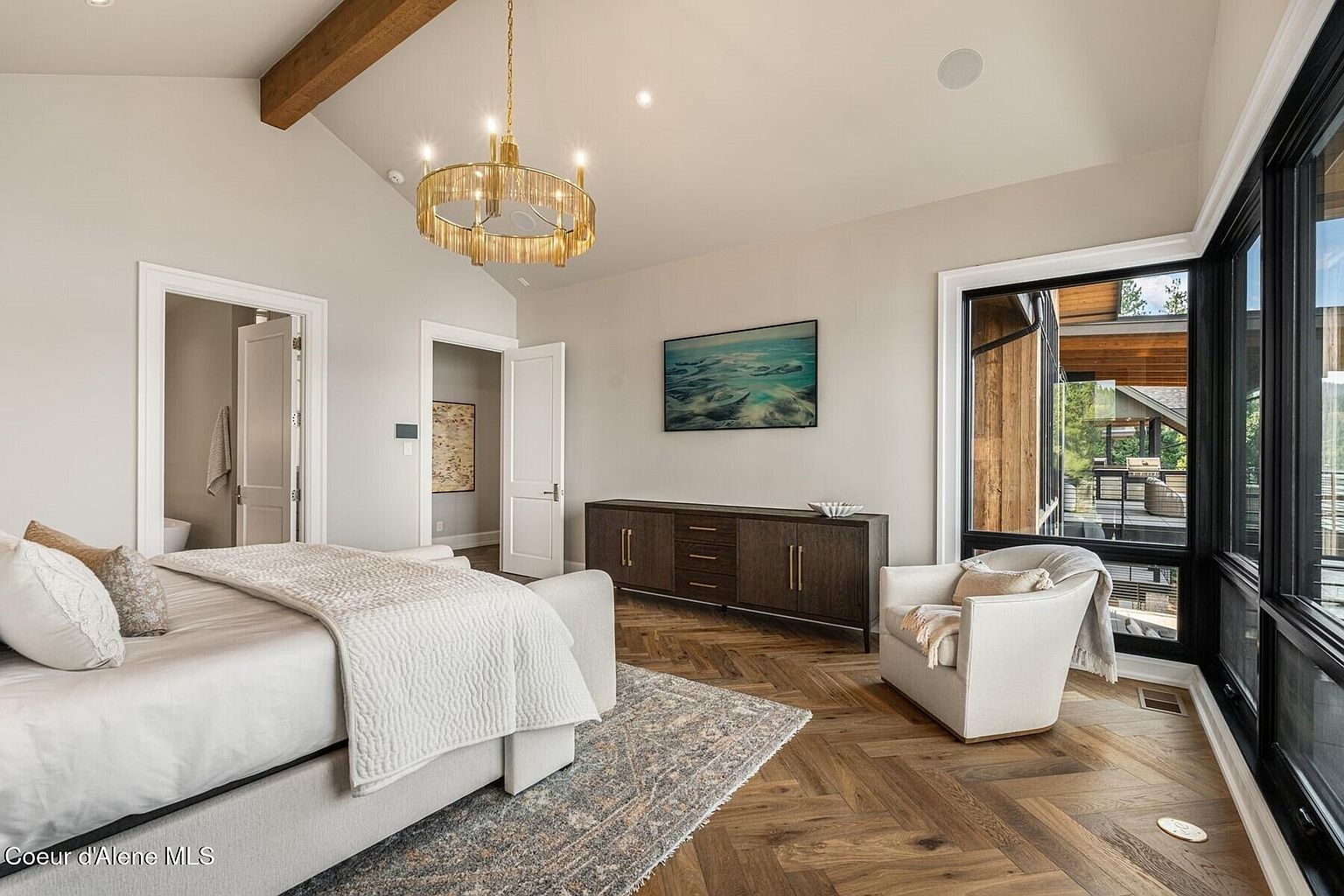 This is a primary bedroom featuring a neutral color palette, hardwood floors laid in a herringbone pattern, and a modern chandelier. The room includes a bed with a light-colored quilt, an armchair near a large window with black frames, and a dark wood credenza with a television mounted above it. The overall impression is one of understated elegance and comfort.