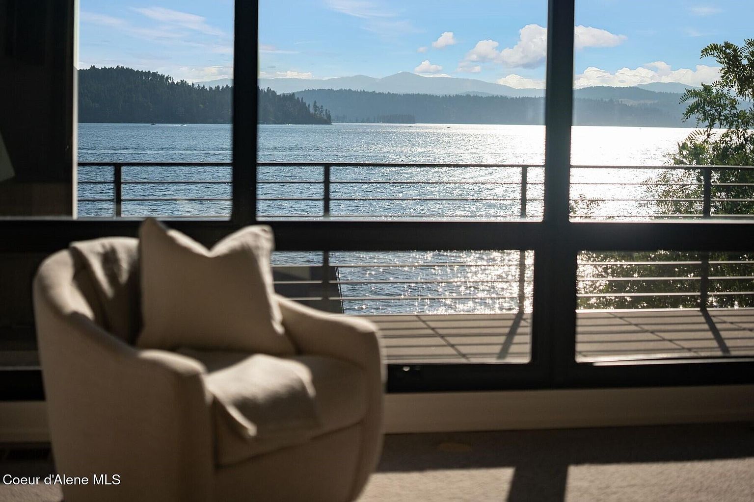 This interior shot showcases a cozy living room with a focus on the stunning lake view through a large window. A comfortable armchair with a plush pillow sits in the foreground, inviting relaxation. The natural light reflecting off the water creates a serene and luxurious atmosphere, perfect for a waterfront property.