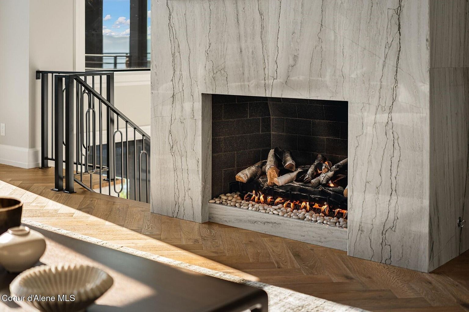This interior shot showcases a modern living room with a striking marble fireplace as the focal point. The fireplace features a dark brick interior and a bed of white pebbles beneath the burning logs. The room also includes a staircase with black metal railings and hardwood floors laid in a herringbone pattern, creating a warm and inviting atmosphere.
