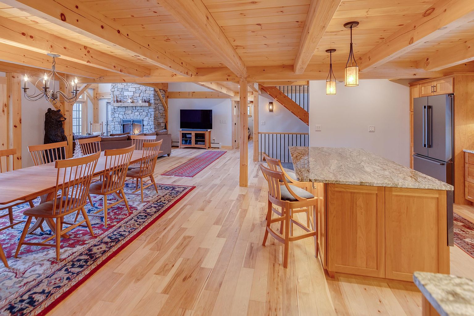 This open-concept living space features a rustic aesthetic with exposed timber beams and light wood flooring throughout. The foreground showcases a kitchen island with a stone countertop and bar seating, while the background reveals a dining area and a living room centered around a stone-clad chimney breast. The perspective is from the kitchen looking toward the living area, highlighting the spacious, interconnected layout of the home.