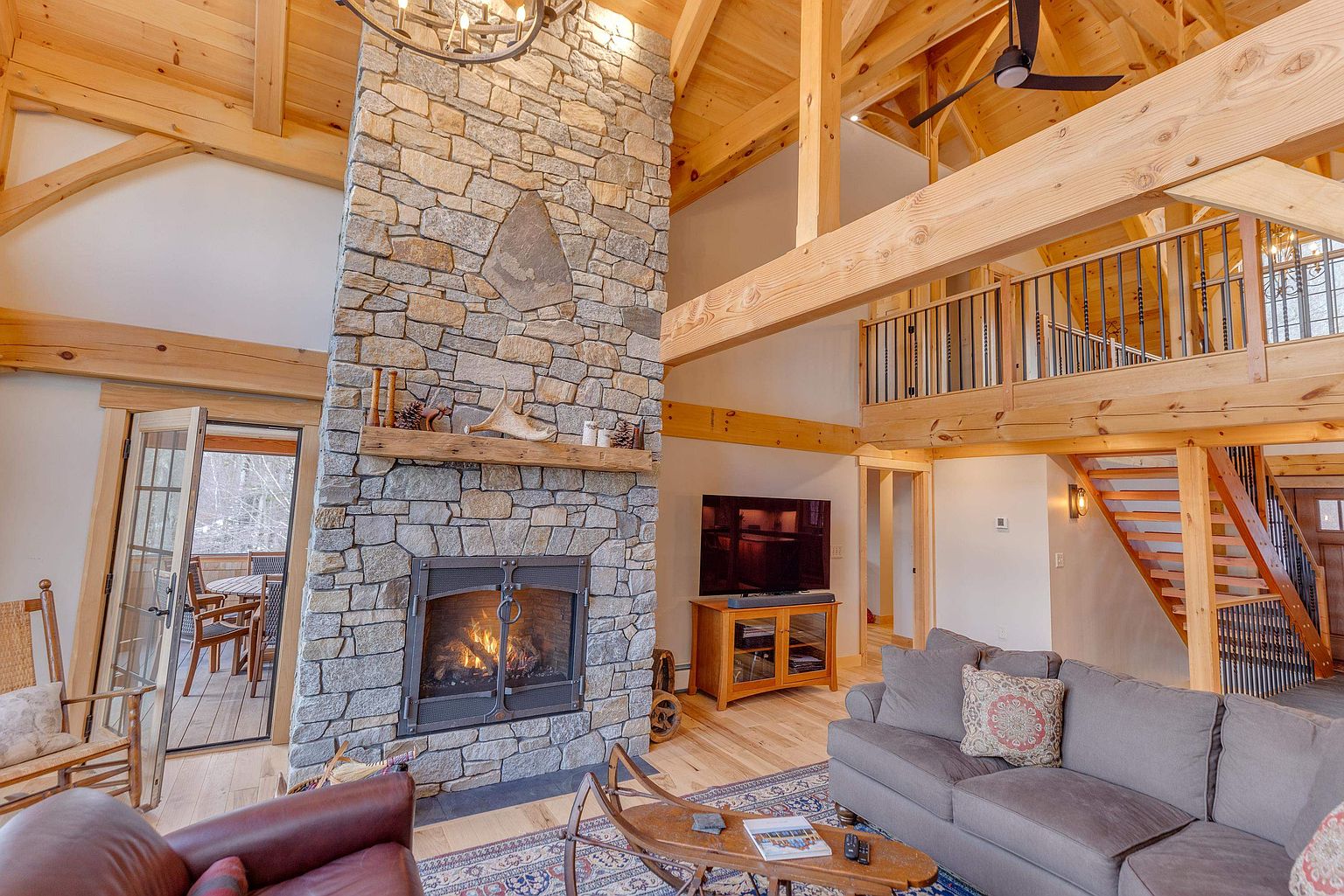 This expansive living room features a rustic aesthetic with exposed timber beams and a prominent stone-clad chimney breast rising above the mantel. The open-concept space includes a cozy seating area with a grey sofa, a wooden coffee table, and a view of an adjacent deck through glass doors. A staircase with metal railings leads to an upper loft area, emphasizing the home's airy, vaulted architecture.