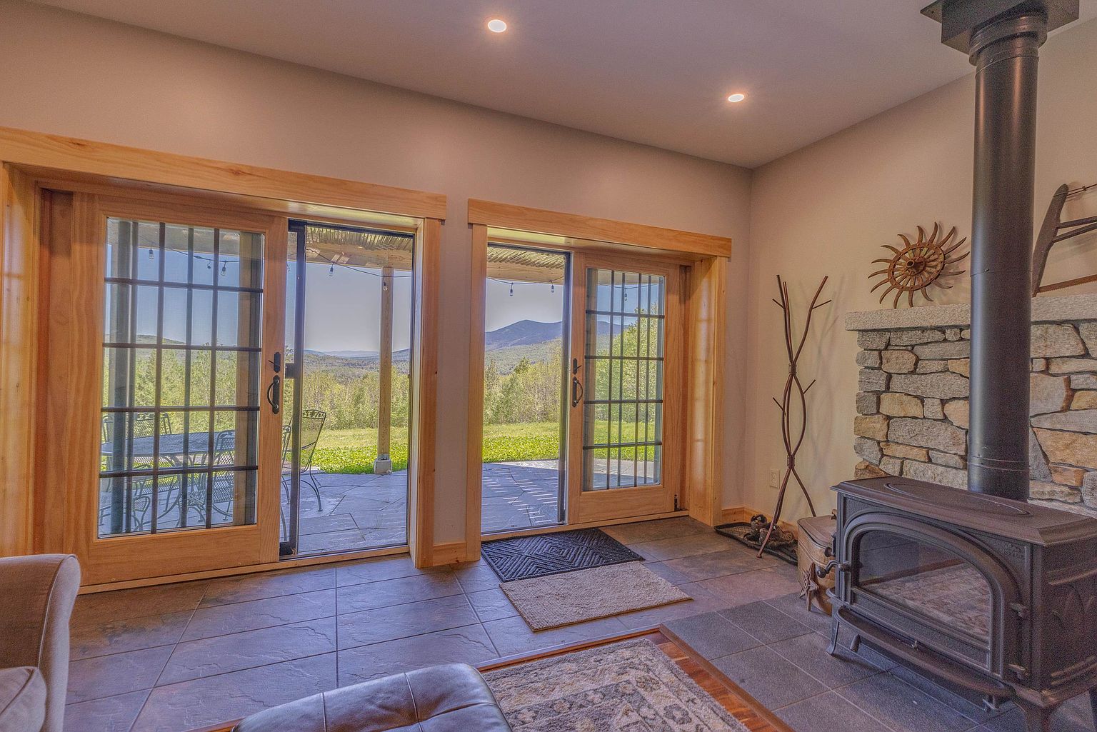 This inviting living space features a rustic wood-burning stove set against a stone-clad chimney breast, creating a cozy focal point. Two sets of sliding glass doors with light wood frames open onto a stone patio, framing expansive views of the surrounding mountains and greenery. The room is finished with dark tile flooring, a patterned area rug, and recessed lighting, offering a warm and comfortable atmosphere.
