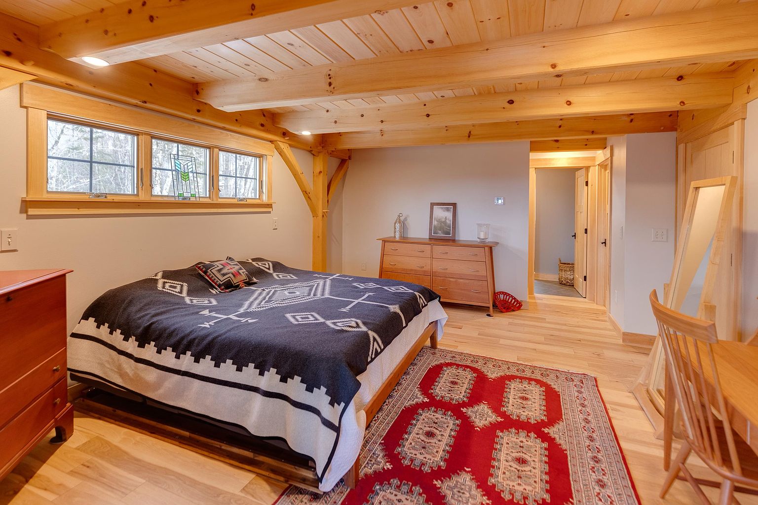 This bedroom features a rustic aesthetic with exposed timber ceiling beams and light wood flooring. A bed with a patterned dark blanket serves as the focal point, complemented by a vibrant red area rug and a wooden dresser against the back wall. The room is brightened by a row of windows that allow natural light to fill the space, creating a warm and inviting atmosphere.
