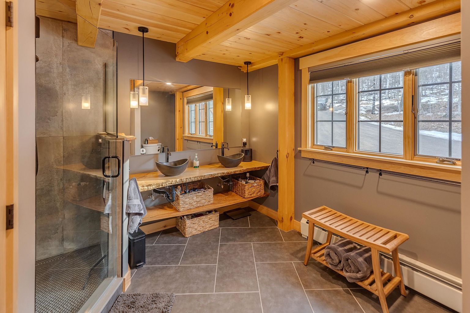 This rustic-inspired bathroom features a double vanity with stone vessel sinks set upon a live-edge wood countertop. The space includes a glass-enclosed shower with gray tile, a wooden bench, and large windows that provide natural light and a view of the outdoors. The combination of exposed wood beams, dark gray walls, and slate-toned floor tiles creates a warm, earthy atmosphere.