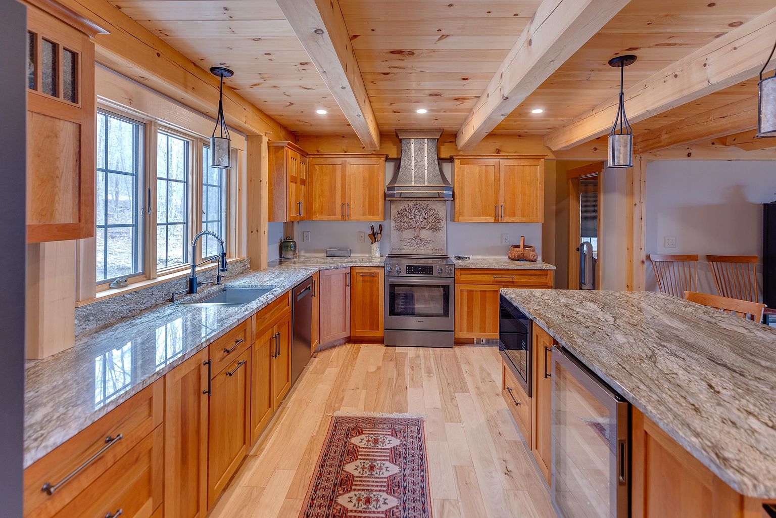 This rustic-inspired kitchen features warm wood cabinetry, exposed ceiling beams, and expansive stone countertops. A central island provides additional workspace and seating, while a decorative tile backsplash behind the stainless steel range adds a unique focal point. The space is illuminated by pendant lighting and natural light from a series of windows above the sink, creating a cozy and inviting atmosphere.