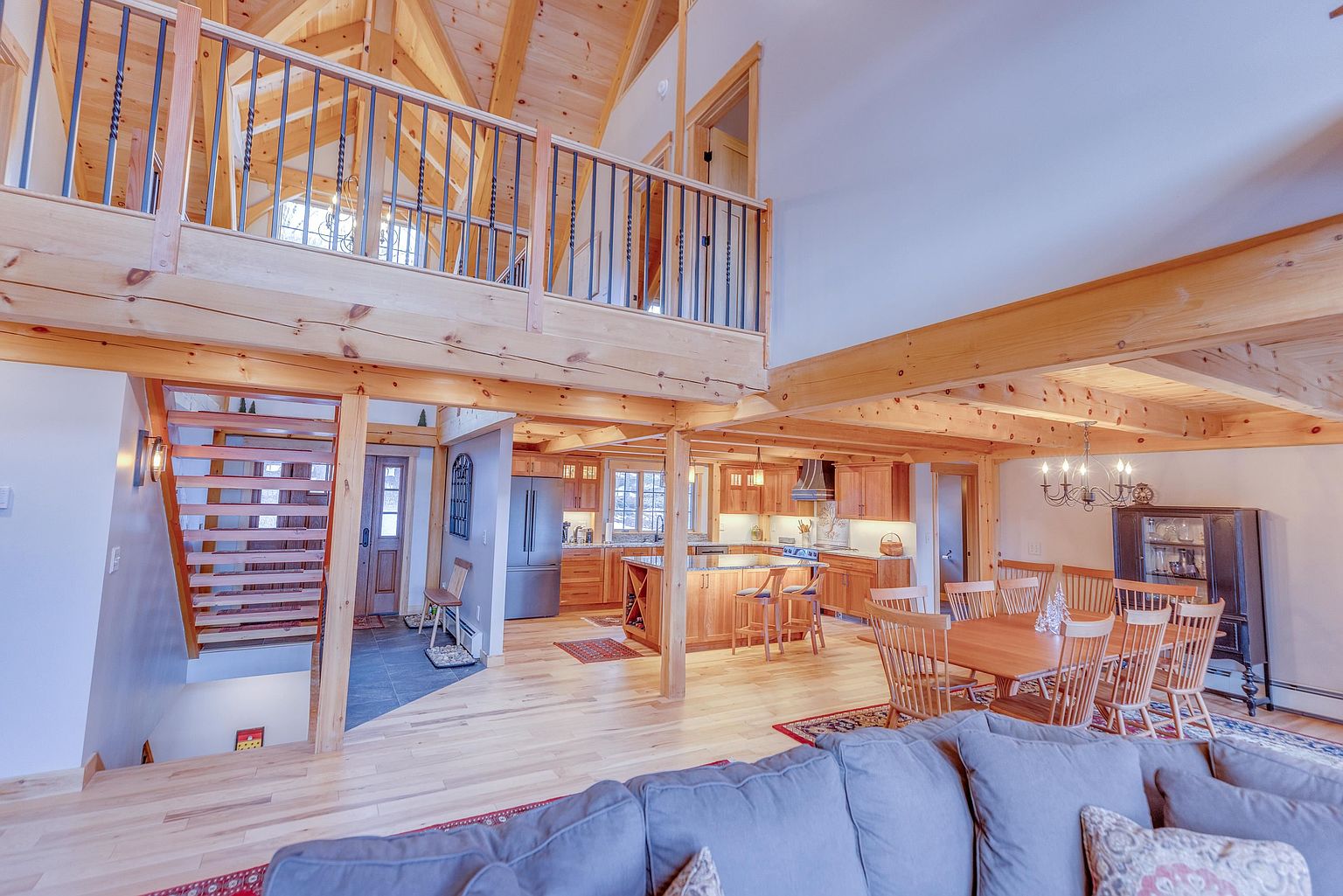 This open-concept living space features a soaring vaulted ceiling with exposed timber beams and a lofted balcony overlooking the main floor. The area seamlessly integrates a dining space with a wooden table, a well-appointed kitchen with a central island, and a cozy living area in the foreground. The warm wood tones, natural light, and rustic architectural details create an inviting and spacious atmosphere.