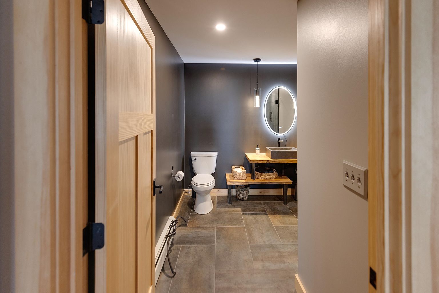 This modern powder room is viewed from a doorway, showcasing a dark, moody color palette contrasted by light wood accents. The space features a floating vanity with a stone vessel sink, a backlit circular mirror, and a minimalist pendant light. Large-format gray tiles cover the floor, leading to a standard toilet positioned against the back wall.