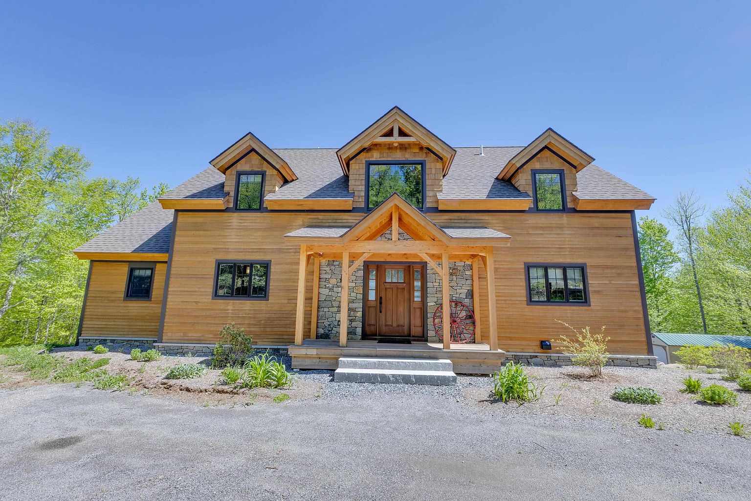 This charming two-story home features a warm, natural wood-sided exterior accented by stone masonry around the entryway and foundation. The front facade is defined by a symmetrical design with three gabled dormers, a covered porch with timber framing, and a central wooden front door. The house is set against a backdrop of lush green trees under a clear blue sky, creating a welcoming and rustic aesthetic.