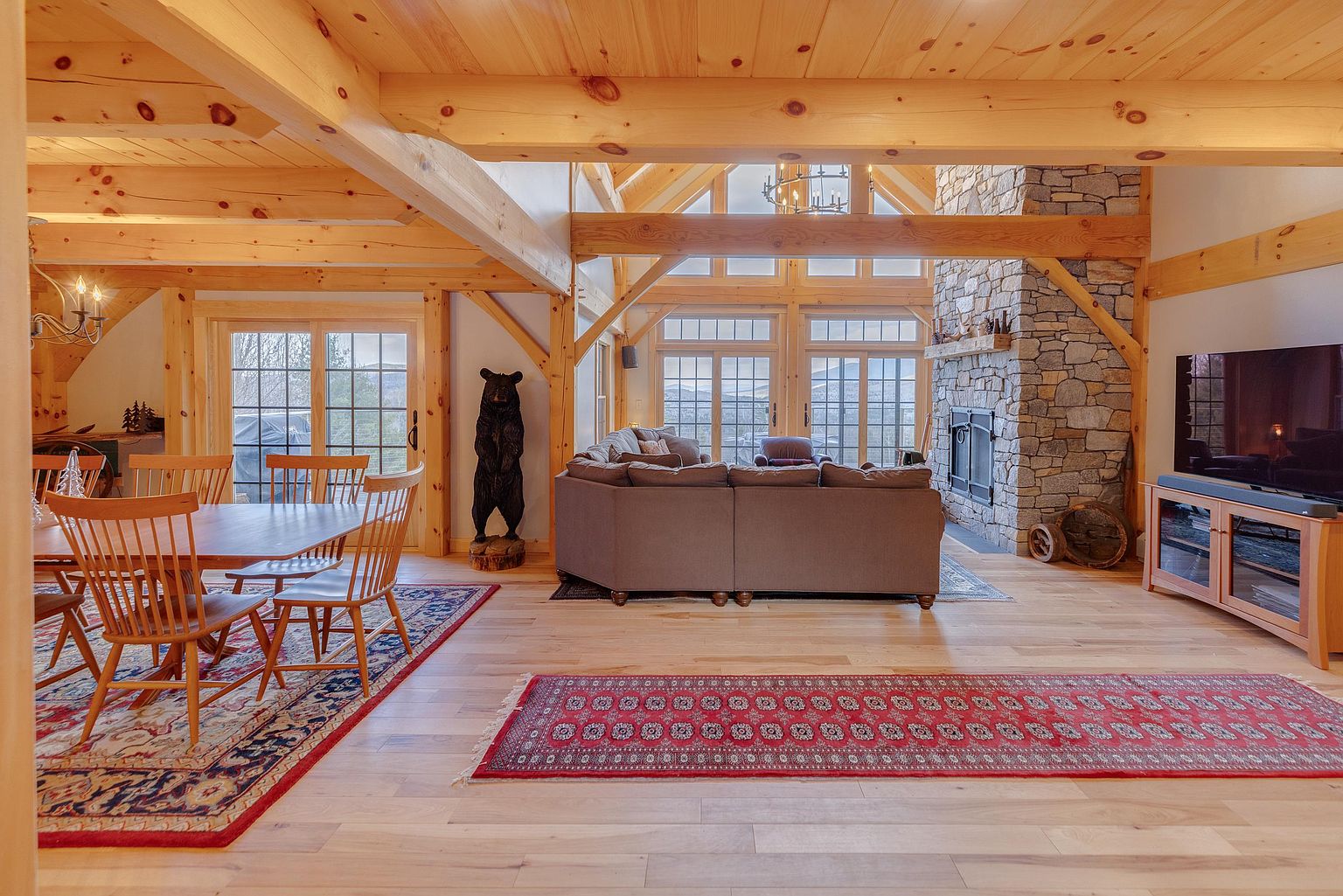 This open-concept living and dining area showcases a rustic timber-frame aesthetic with exposed wooden beams and light wood flooring. A prominent stone-clad chimney breast serves as a focal point near a large sectional sofa, while a dining set sits on a patterned rug in the foreground. The space is bathed in natural light from large windows that offer expansive views, creating a warm and inviting atmosphere.
