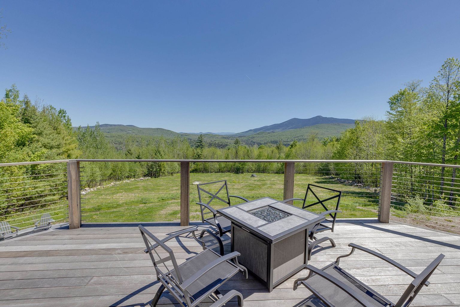 This elevated wooden deck offers a panoramic view of lush green mountains and a sprawling landscape under a clear blue sky. The space is furnished with a square fire pit table surrounded by four metal-framed chairs, creating an inviting outdoor seating area. A modern cable railing system provides safety while maintaining an unobstructed view of the scenic surroundings.