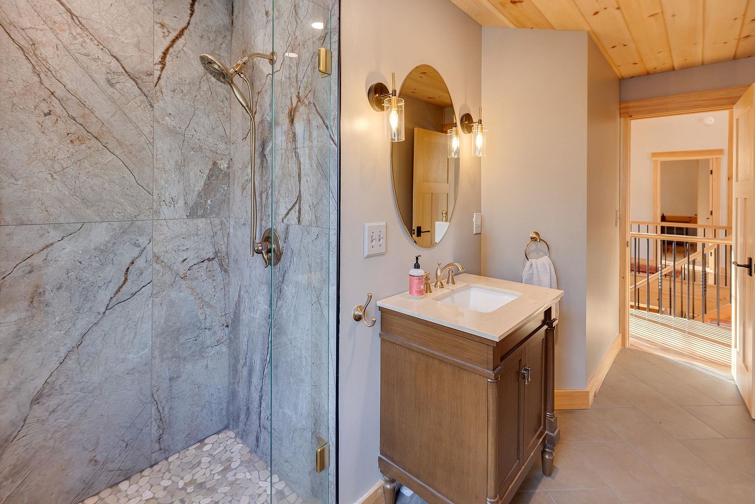 This bathroom features a walk-in shower with large-format stone-look wall tiles and a pebble-textured floor, enclosed by a glass panel. A wooden vanity with a light-colored stone top sits adjacent to the shower, complemented by a round mirror and dual wall-mounted light fixtures. The space includes a neutral color palette, wood-paneled ceiling accents, and a view into an adjacent hallway with a staircase.