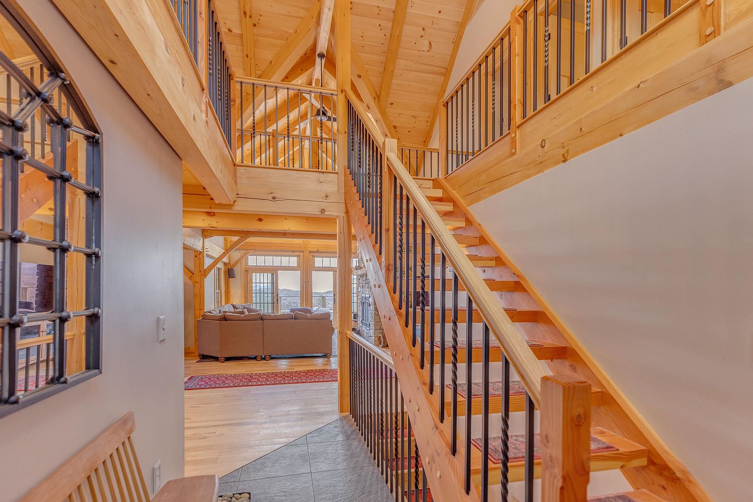This perspective captures a grand entryway featuring a prominent wooden staircase with black metal balusters leading to an upper landing. The open-concept design reveals a living area in the background, characterized by light wood beams, vaulted ceilings, and large windows that invite natural light. The space exudes a rustic, lodge-like aesthetic with warm wood tones and a clean, inviting atmosphere.